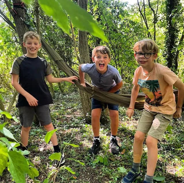 Three young boys playing in a forest with a hammock, two smiling and one with an open mouth expression.