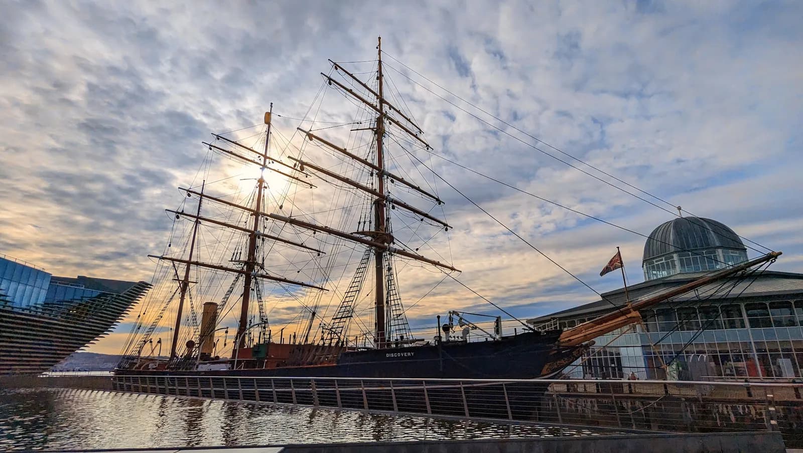 A large sailing ship with multiple tall masts and rigging docked in front of a modern building with a distinct architectural design and another building with a domed roof, all under a cloudy sky.