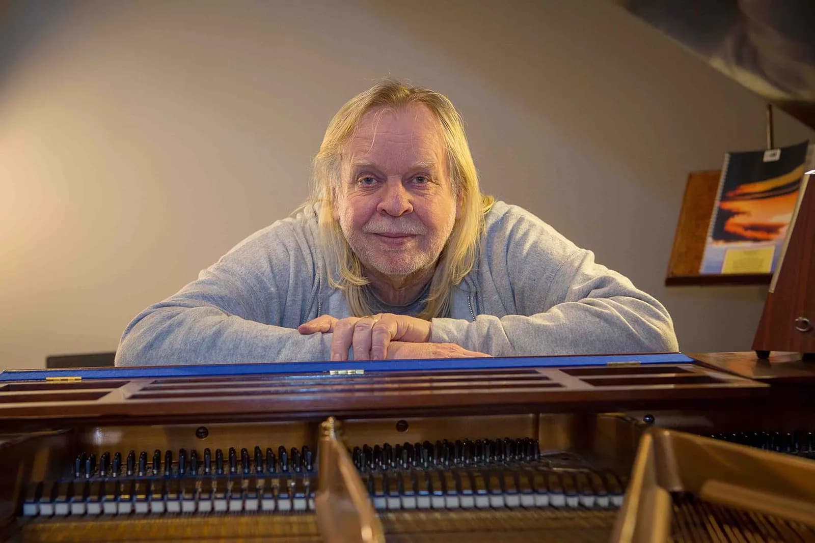 A man with long blonde hair and a beard, wearing a grey sweatshirt, leans over an open piano and looks directly at the camera.