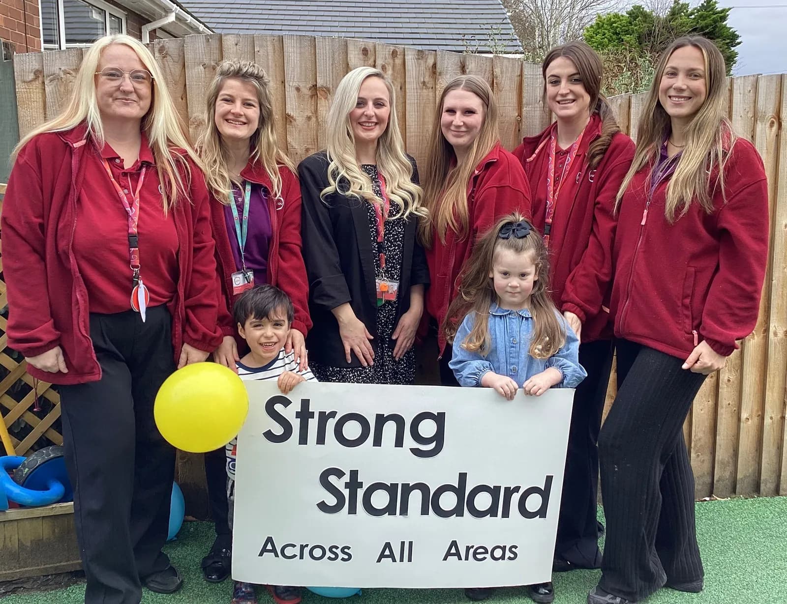 Six women and two young children pose for a photo in front of a wooden fence, with the children holding a sign that reads "Strong Standard Across All Areas."