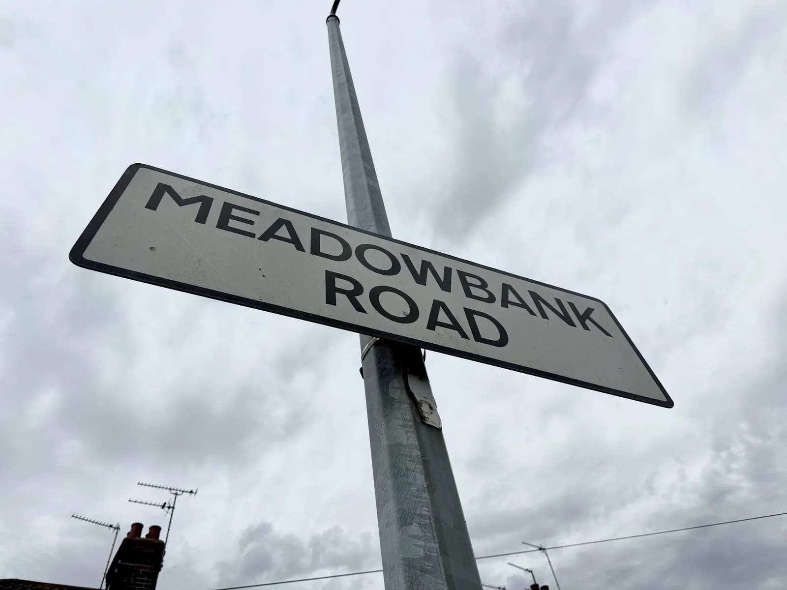A street sign for Meadowbank Road is angled upwards against a cloudy sky, with parts of a chimney and antenna visible in the bottom left.