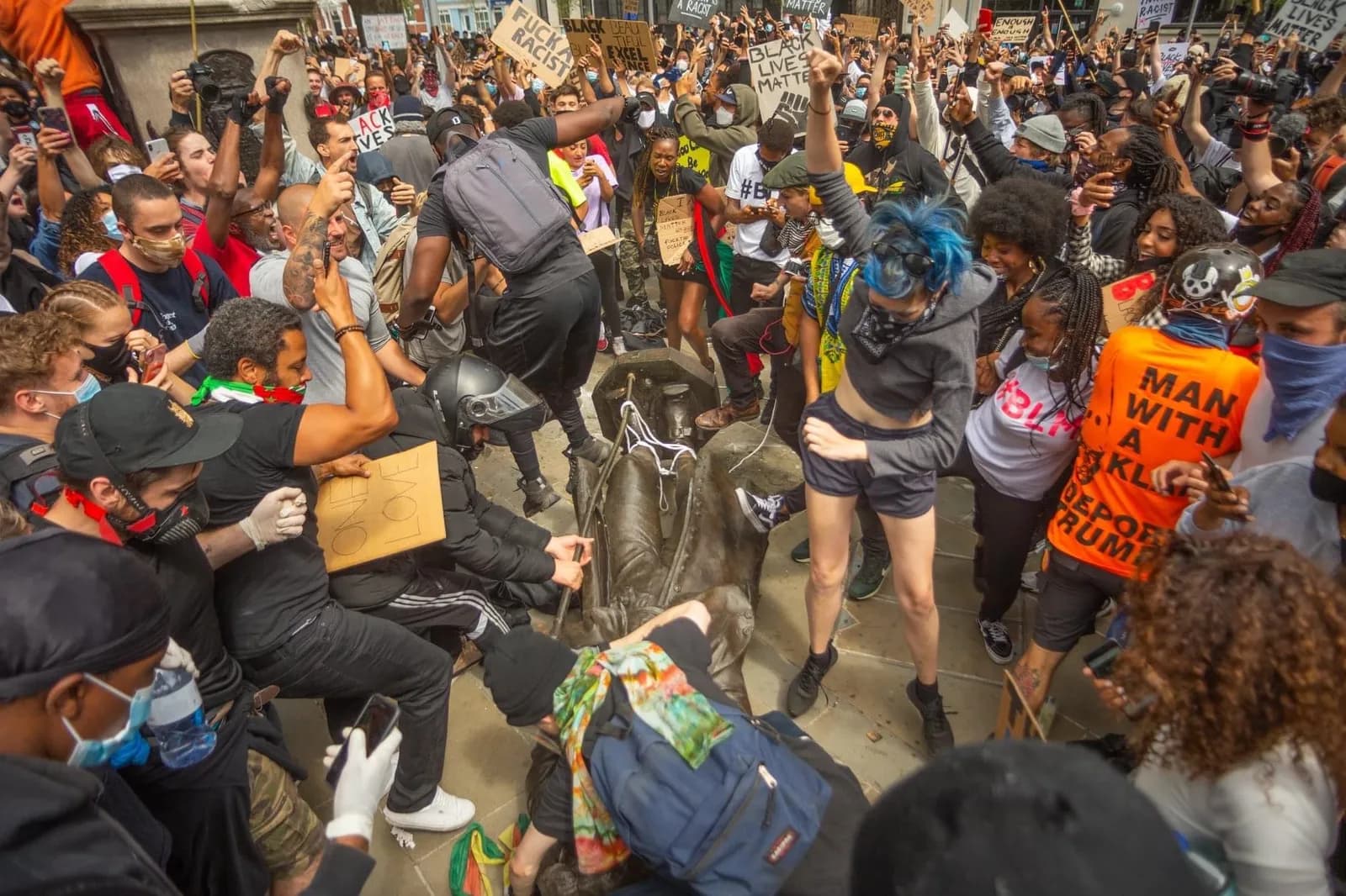 A diverse crowd of protesters surrounds and climbs on a bronze statue, with some people holding signs and raising their fists in the air.