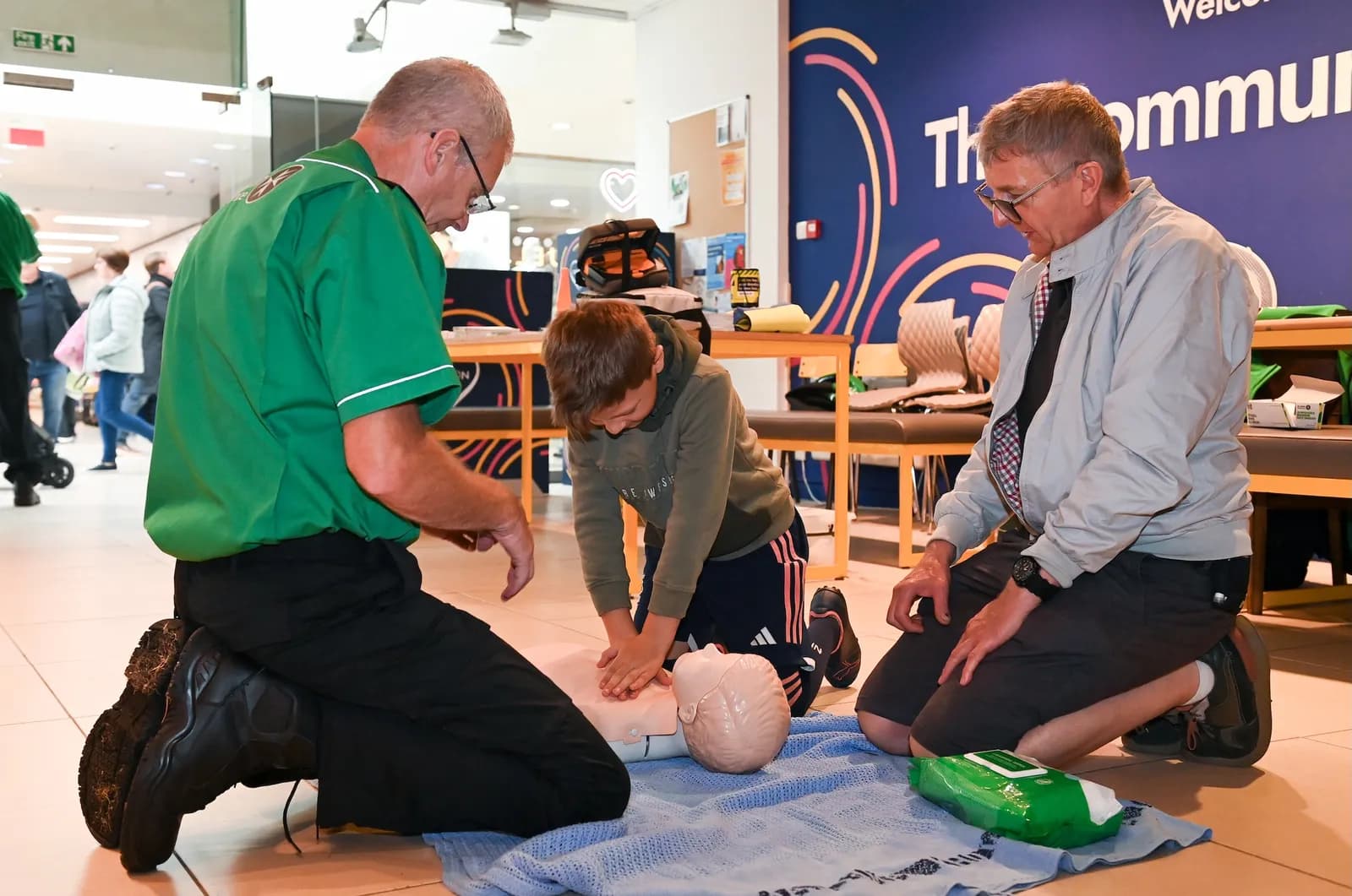 A young boy practices CPR on a mannequin while two adult men, one in a green shirt and one in a grey jacket, kneel beside him, observing.