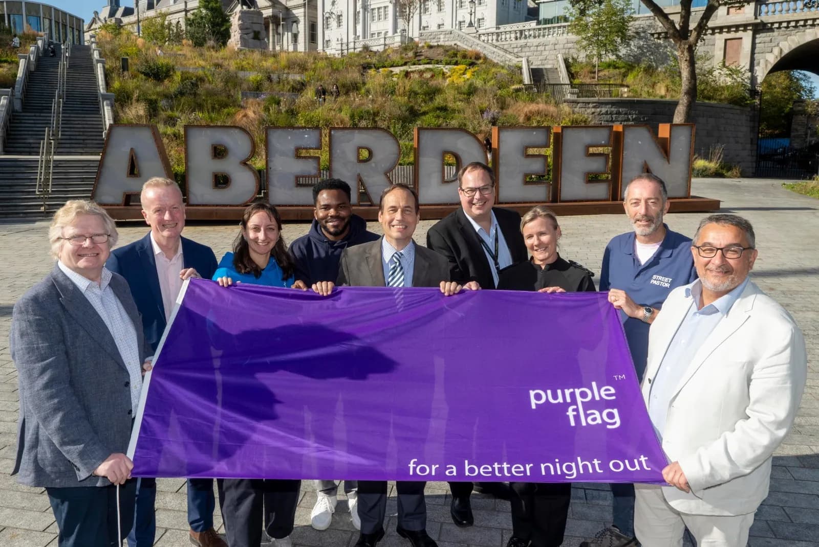A group of nine diverse adults hold a large purple banner that reads "purple flag for a better night out" in front of a giant "ABERDEEN" sign.