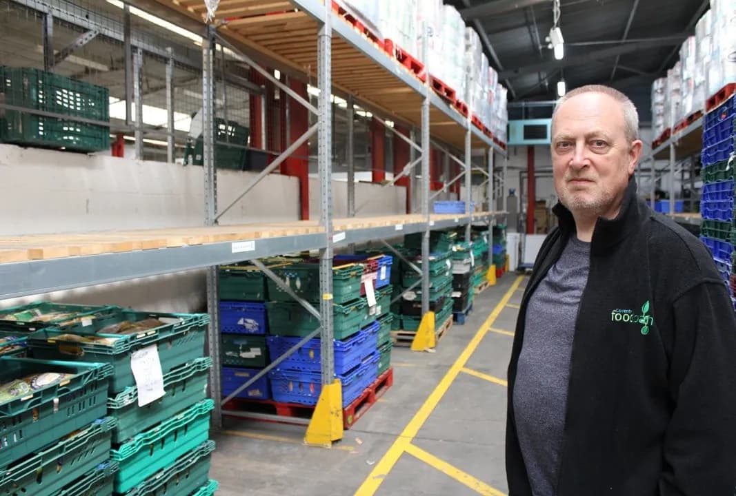 A man in a black jacket and grey t-shirt stands amidst warehouse shelving filled with stacks of green and blue crates.