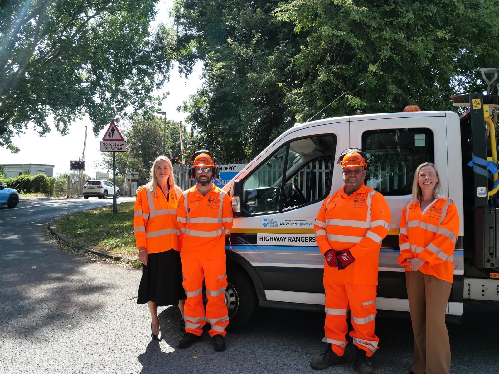 Four highway rangers in bright orange vests and two in hard hats stand next to a white Highway Rangers van on a sunny day.