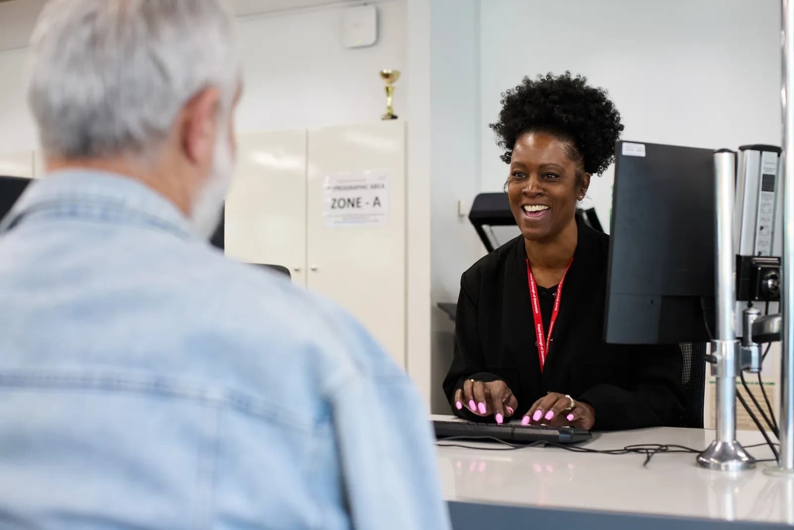 A smiling woman with dark curly hair and a red lanyard around her neck sits at a desk typing on a computer, looking towards an older man with grey hair whose back is to the viewer.