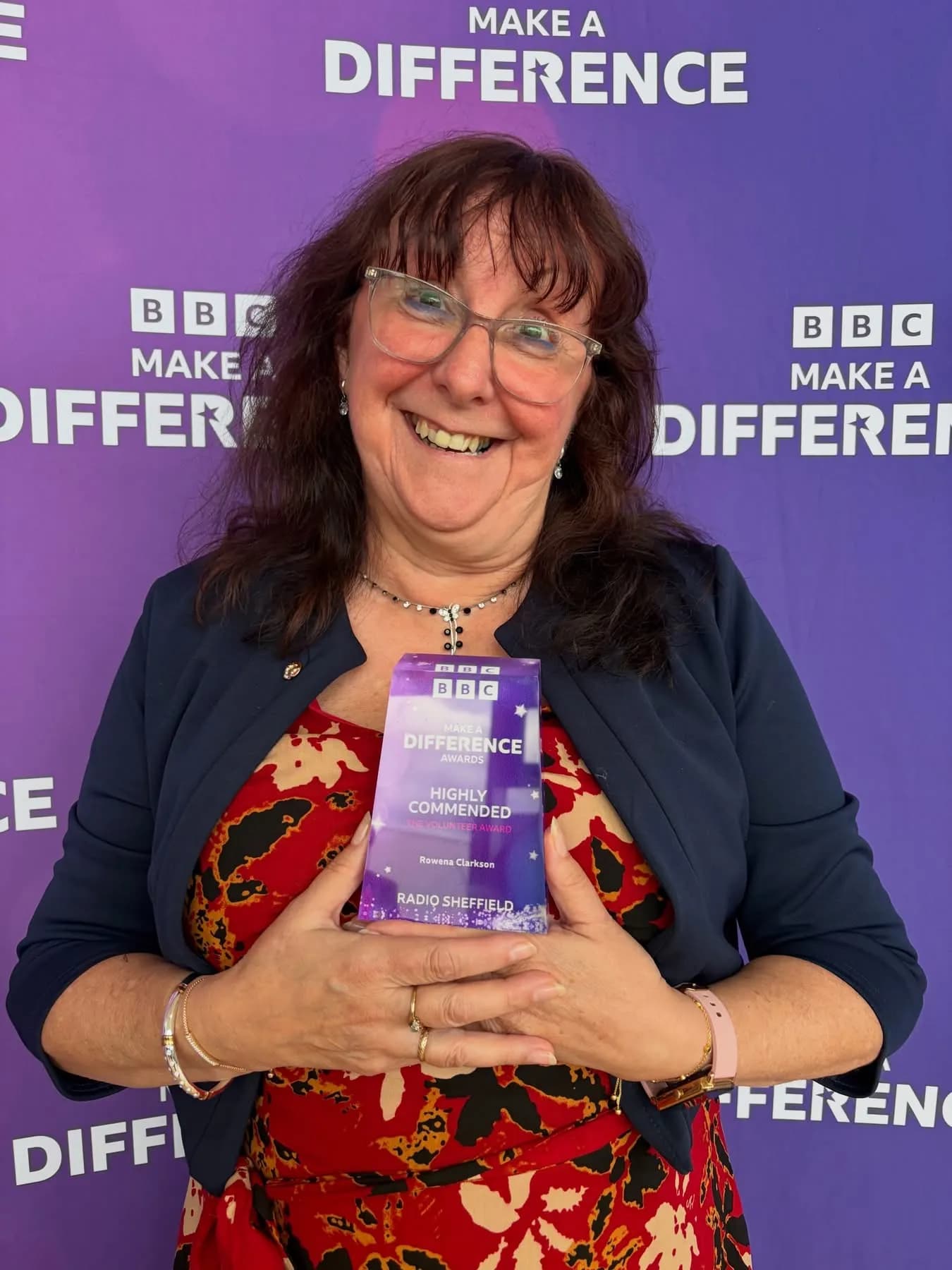 A smiling woman with glasses and long brown hair holds a purple and white award trophy that reads "BBC Make a Difference Awards, Highly Commended, Rowena Clarkson, Radio Sheffield."