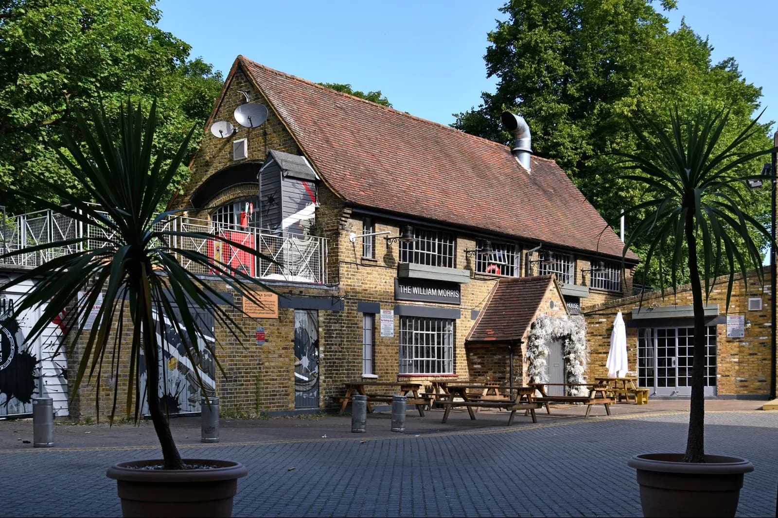 The exterior of The William Morris pub, a brick building with a red tile roof, surrounded by trees and two potted plants.