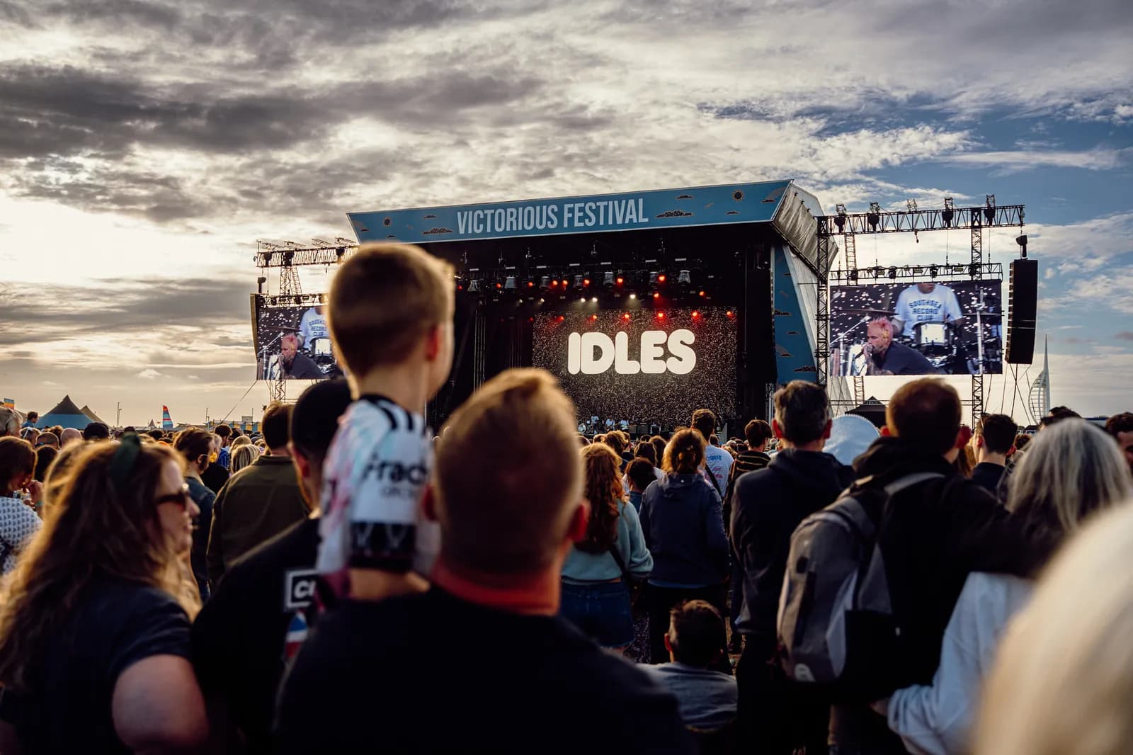 A crowd of people watches the band Idles perform on an outdoor stage at the Victorious Festival as the sun sets.