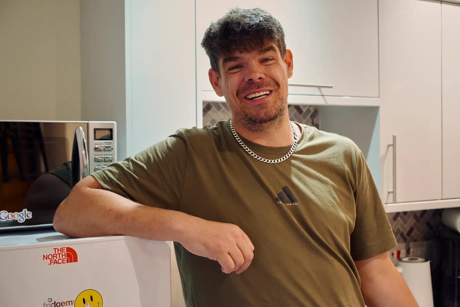 A man with dark, curly hair and a beard, wearing a green t-shirt and silver chain, smiles broadly while leaning on a white appliance in a kitchen.
