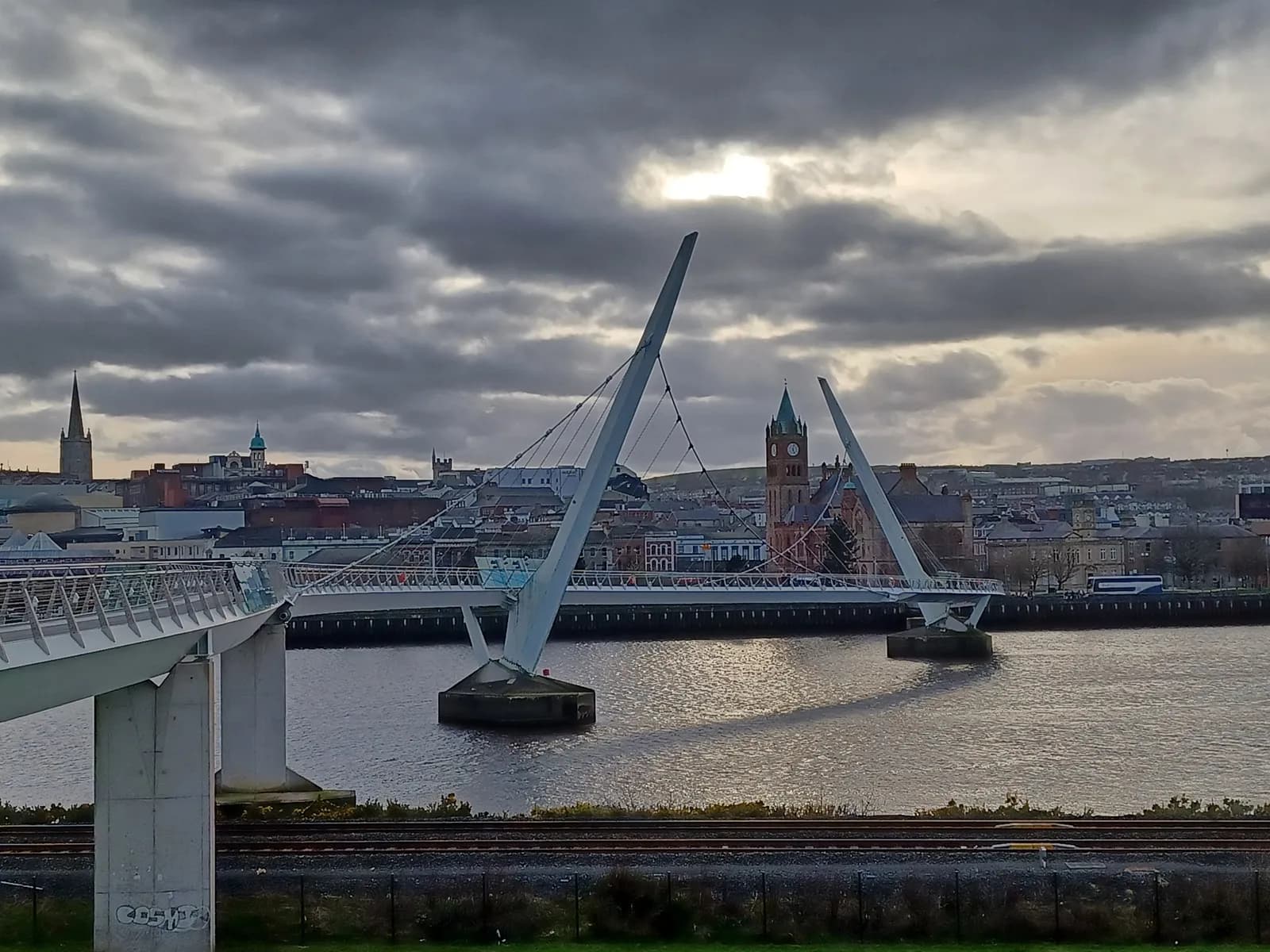 The Peace Bridge curving over water, connecting two sides of a city with historical buildings and a prominent clock tower under a cloudy sky.