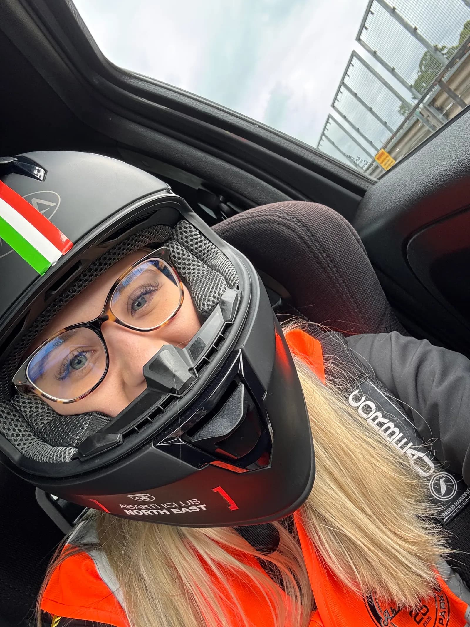 A blond young woman wearing a black helmet, glasses, and an orange safety vest, looks slightly up and to the right.
