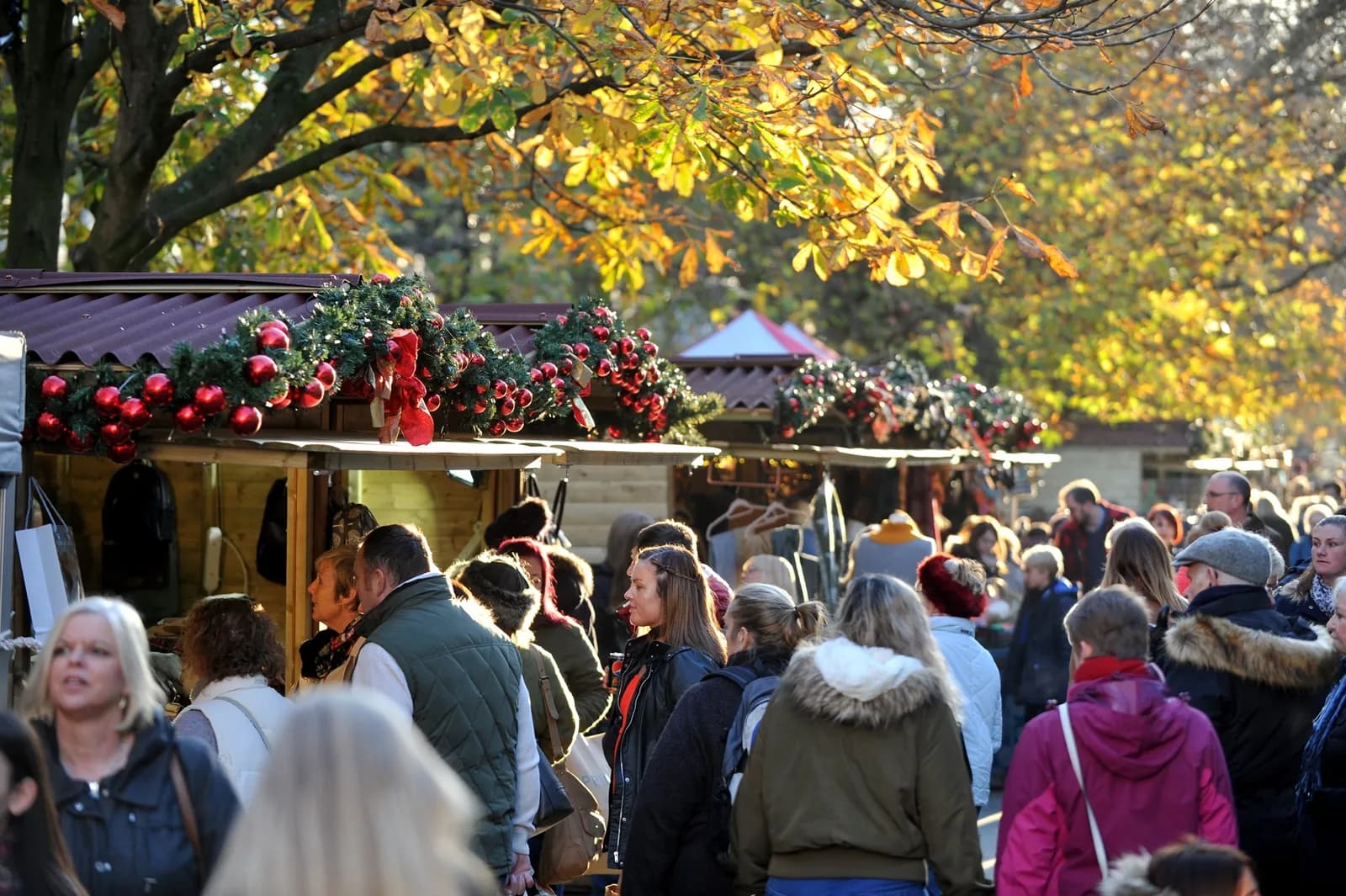 A busy outdoor Christmas market with stalls decorated with red baubles and garlands, and numerous people browsing under trees with autumn leaves.