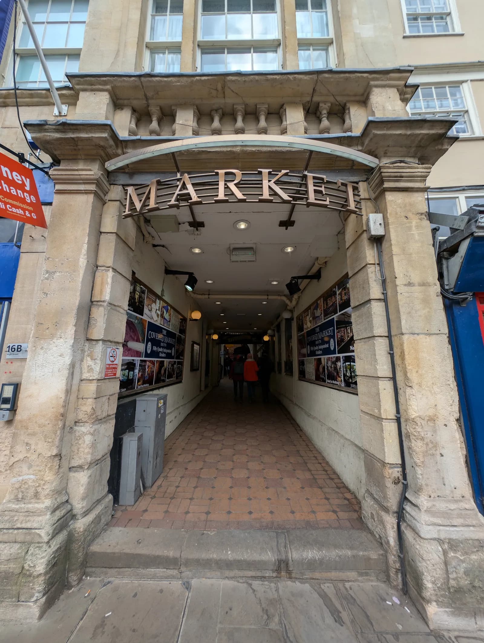 The entrance to a covered market, with the word "MARKET" displayed prominently above a stone archway, leading into a long corridor with tiled flooring and people walking in the distance.