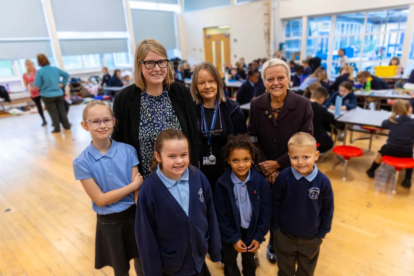 Three adult women and four children in school uniforms smile at the camera while standing in a brightly lit school hall with other students and tables in the background.