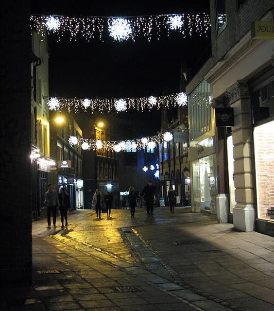 A nighttime view of a street decorated with snowflake-shaped lights strung overhead, with people walking on the wet pavement reflecting the light from shops and streetlights.