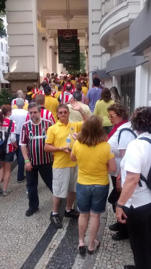 A crowd of people in red, white, yellow, and blue shirts gathered on a Portuguese mosaic sidewalk, with a man in a yellow shirt holding a water bottle and waving his hand in the lower right.