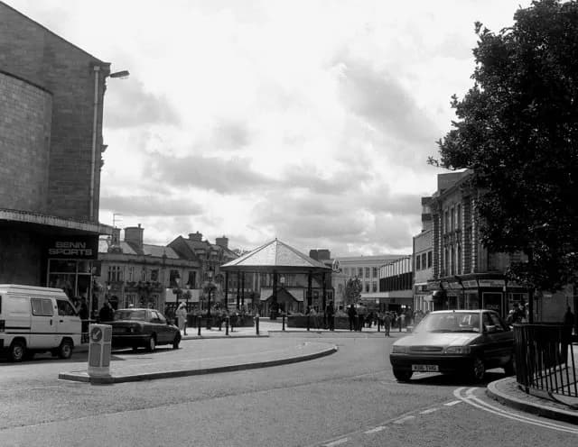 A black and white street scene with buildings flanking a circular road around a central gazebo, with cars parked and driving, and people walking.