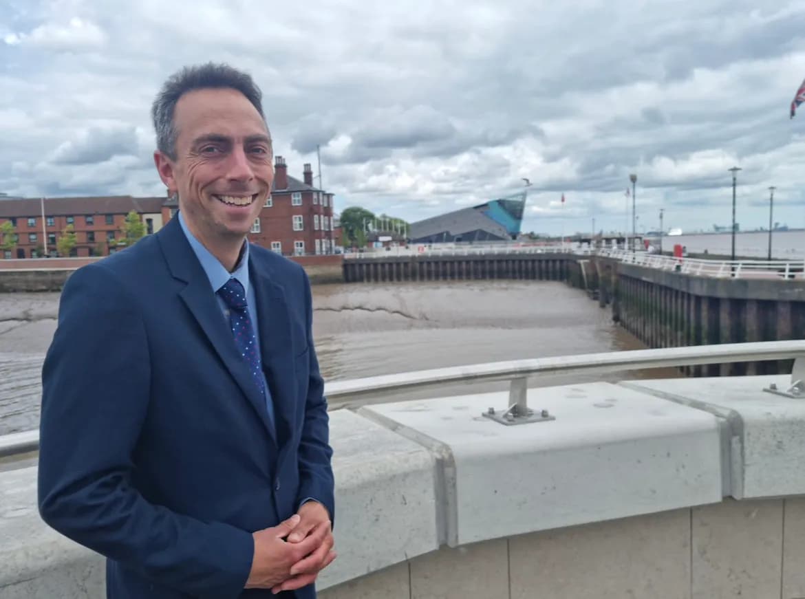 A man in a suit smiles at the viewer, standing on a bridge over a muddy river with buildings in the background under a cloudy sky.