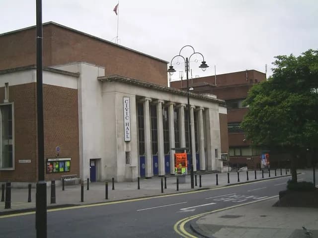 The exterior of a beige Civic Hall building with columns and a sign, next to a street with a lamp post and some trees.
