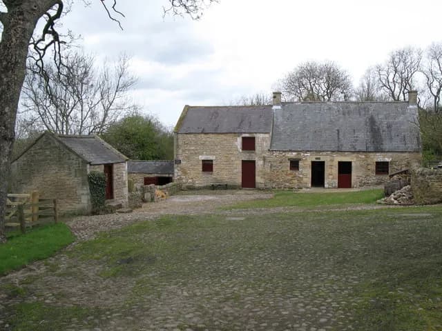 A rustic stone farmhouse and an outbuilding with red doors, surrounded by green grass and a large tree, under a cloudy sky.