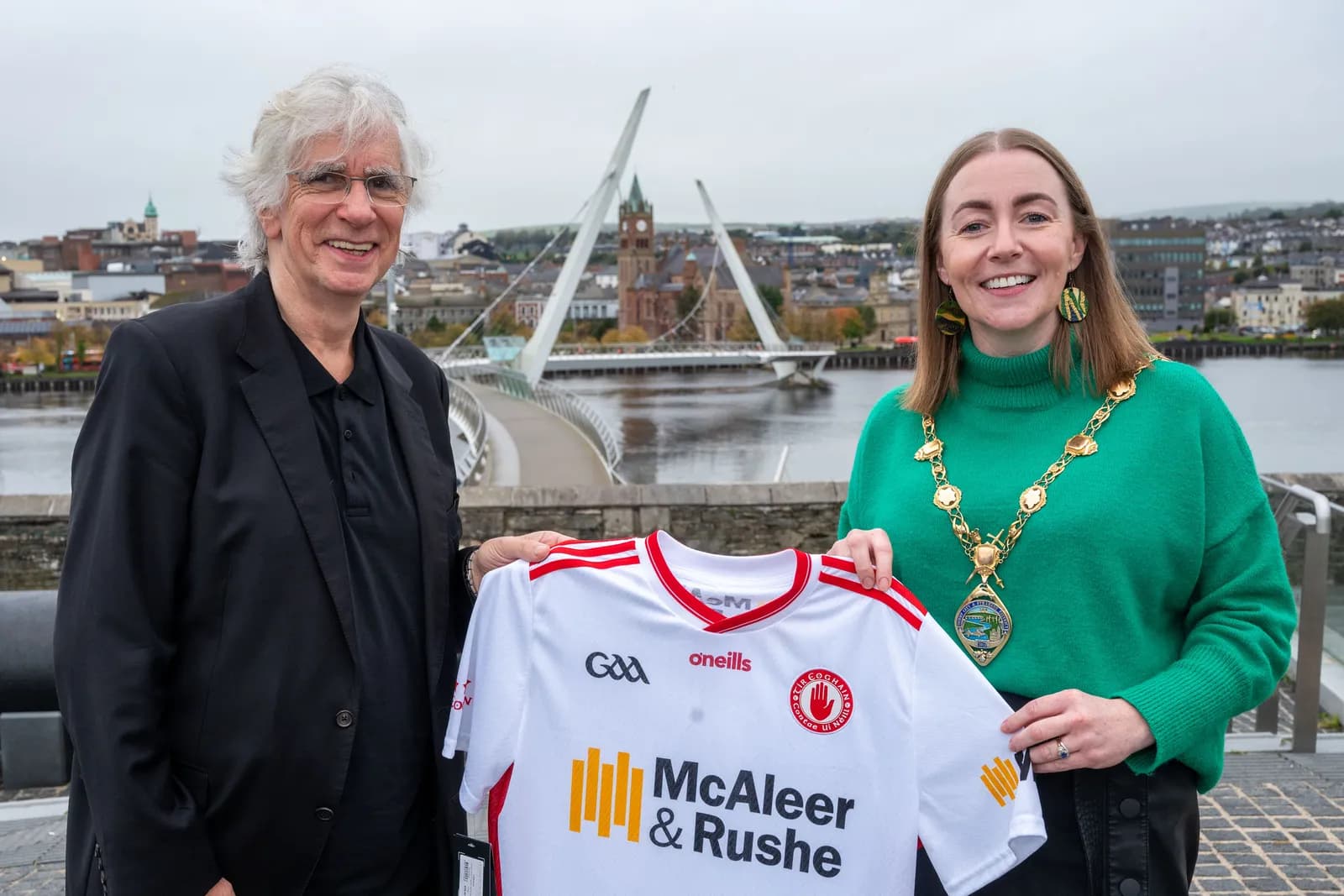 A man with grey hair and glasses in a black suit and a woman in a green sweater with a gold chain are holding up a white and red athletic jersey, with a cityscape and a bridge in the background.