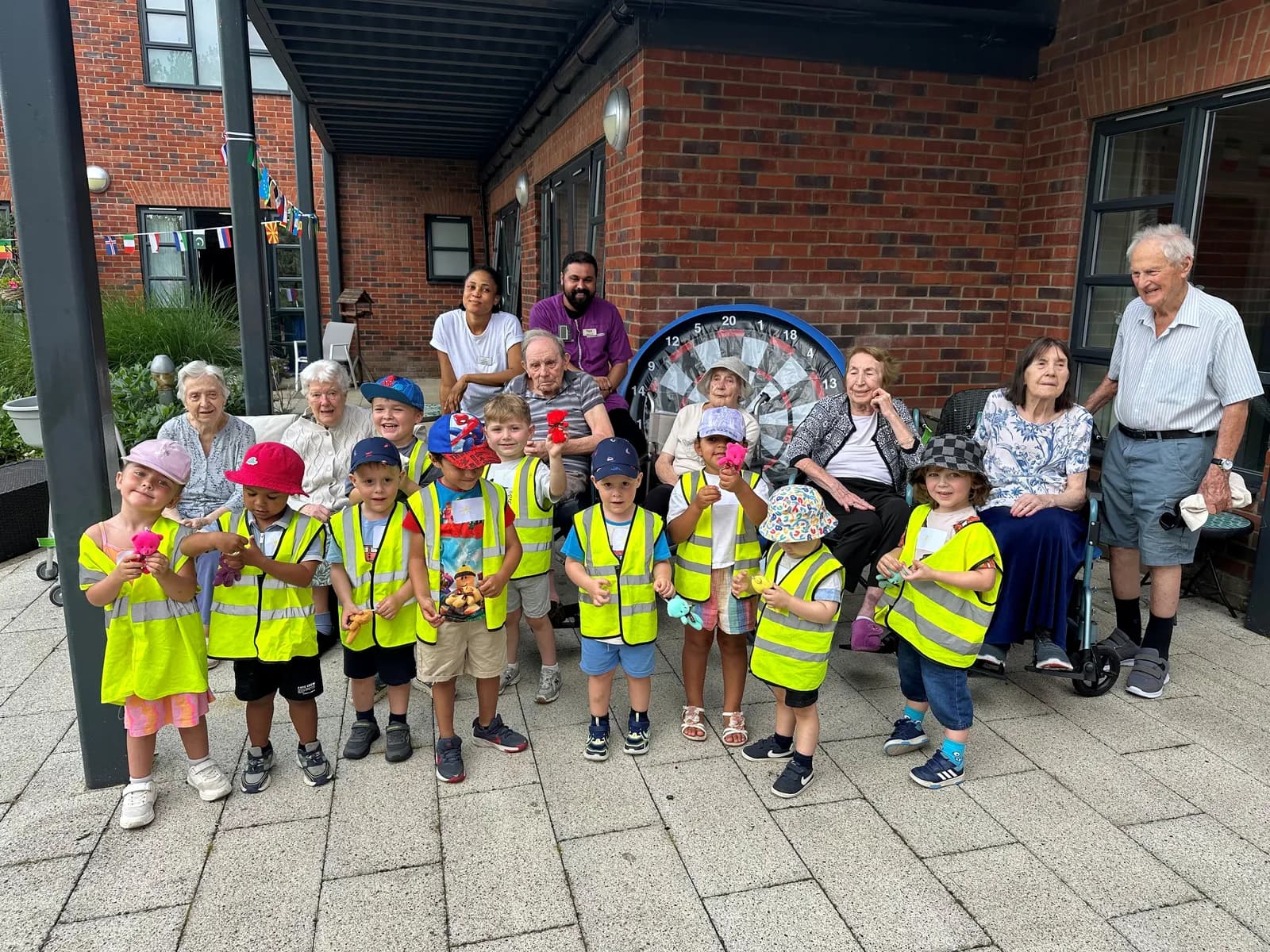 A group of young children wearing high-visibility vests posing with elderly adults and caregivers in front of a brick building with a dartboard.