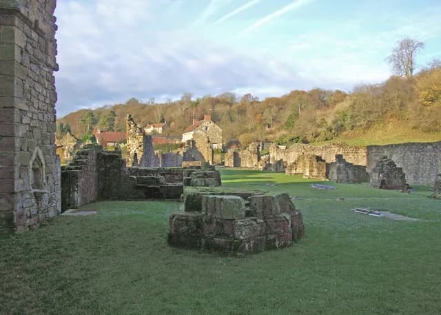 A scenic view of ancient stone ruins on a grassy field with a hillside of trees and houses in the background under a blue sky.