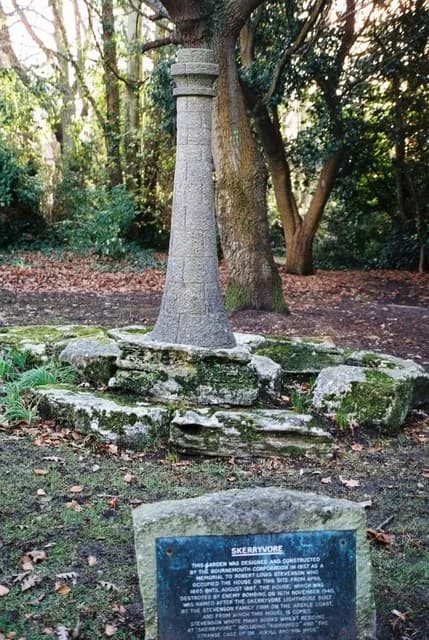A stone monument in a park setting, surrounded by trees and a commemorative plaque on a rock.