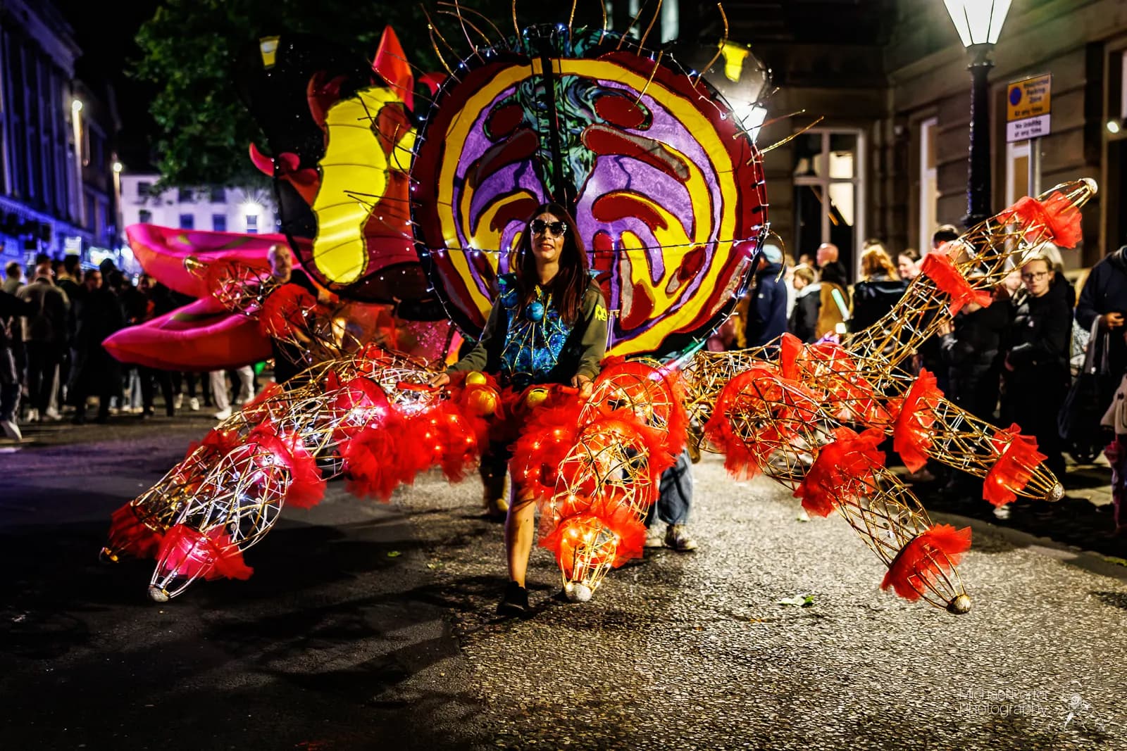 A person wearing a large, colorful, illuminated costume resembling a mythical creature or insect with prominent red, yellow, and purple elements and glowing red lights, walking down a street at night with a crowd of people in the background.