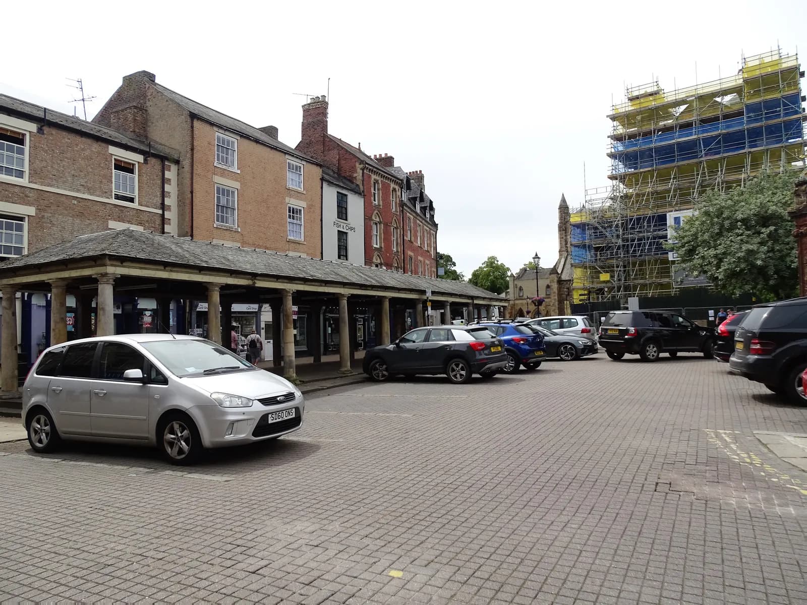 A silver car is parked in a cobblestone lot next to a historic building with pillars, while other cars are parked further back and a large building covered in scaffolding is visible in the background.