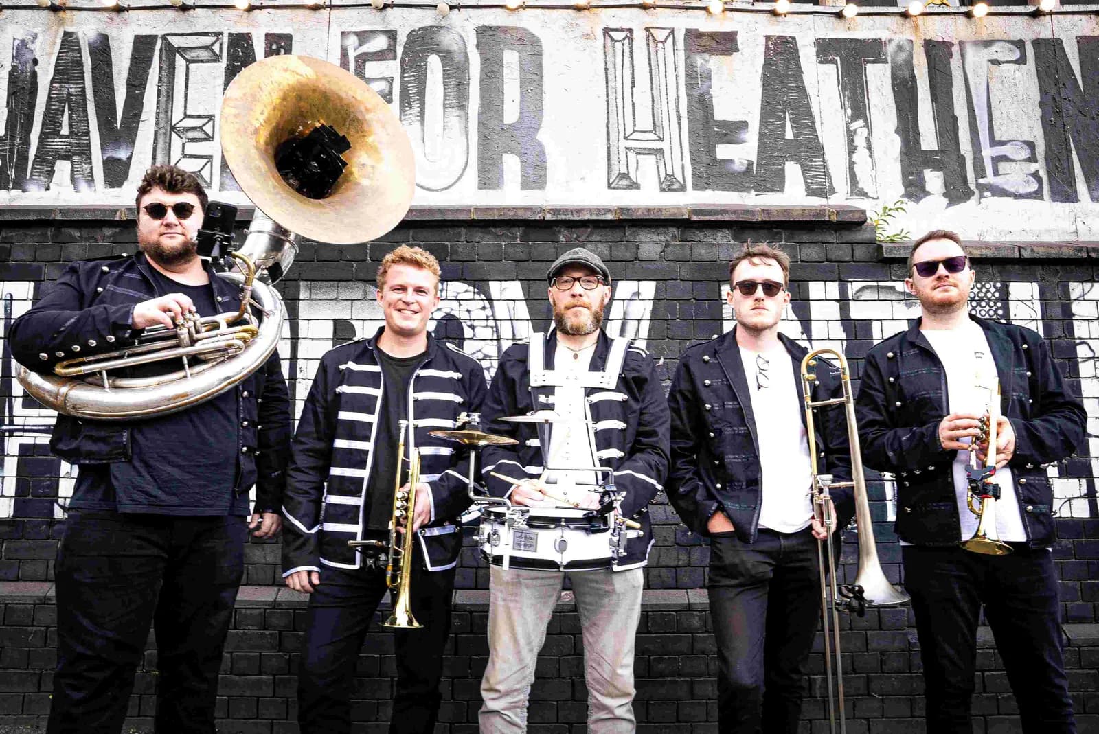 A brass band of five men stands in front of a brick wall with white graffiti on it, all wearing dark jackets with white stripes and sunglasses, holding musical instruments.