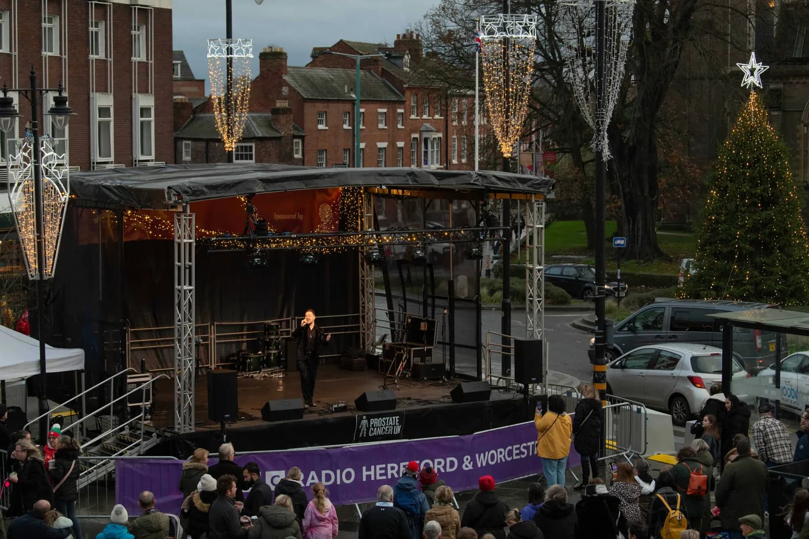 A male singer performing on an outdoor stage at dusk, illuminated by lights, with an audience gathered in front of him.