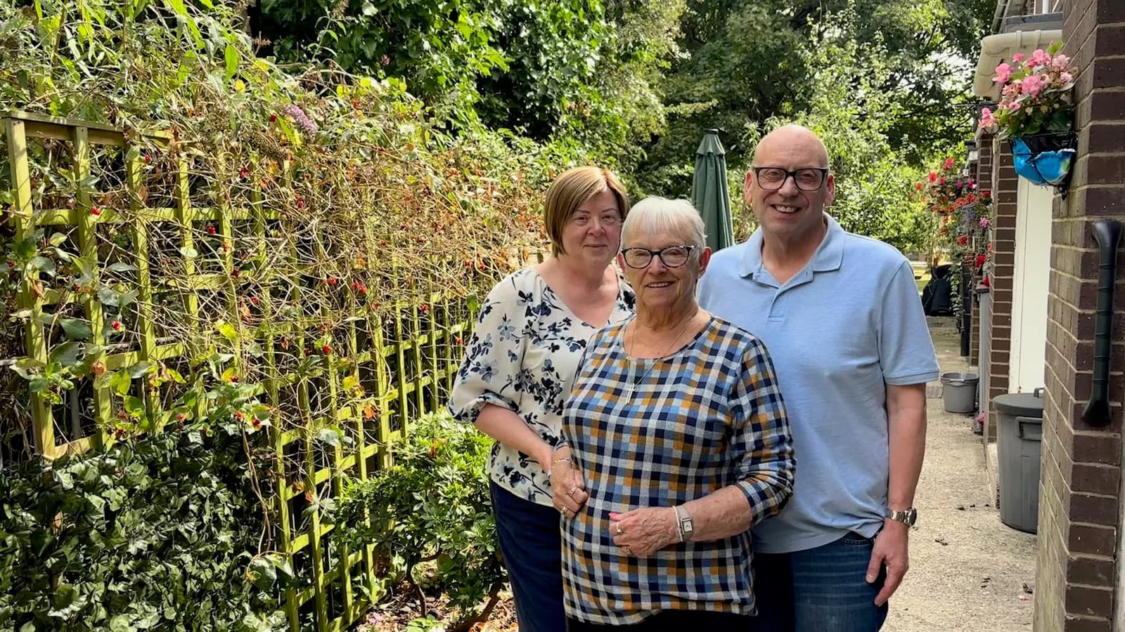 Three individuals, two women and one man, standing together in an outdoor setting with green foliage and a wooden trellis behind them, looking at the camera.