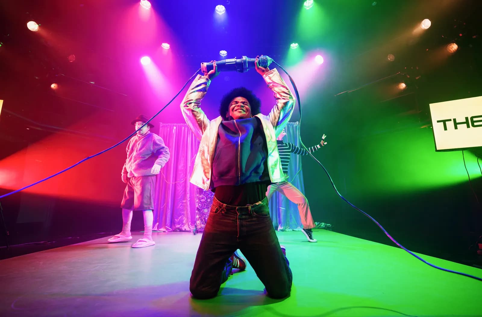 A man with dark curly hair and a shiny gold jacket is on his knees on a stage, holding a long black cable over his head, with two other people and stage lights in the background.