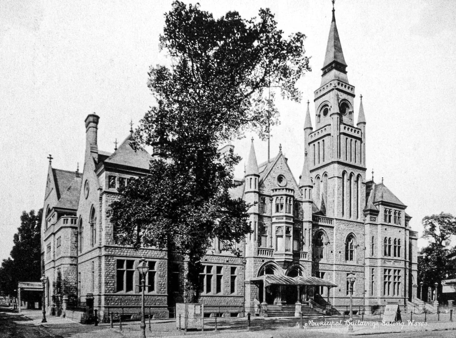 A black and white photograph captures the grandeur of the Municipal Buildings in Swansea, Wales, a large stone structure featuring a prominent clock tower with a spire, ornate architectural details, and several arched windows, partially obscured by a large, leafy tree on the left.