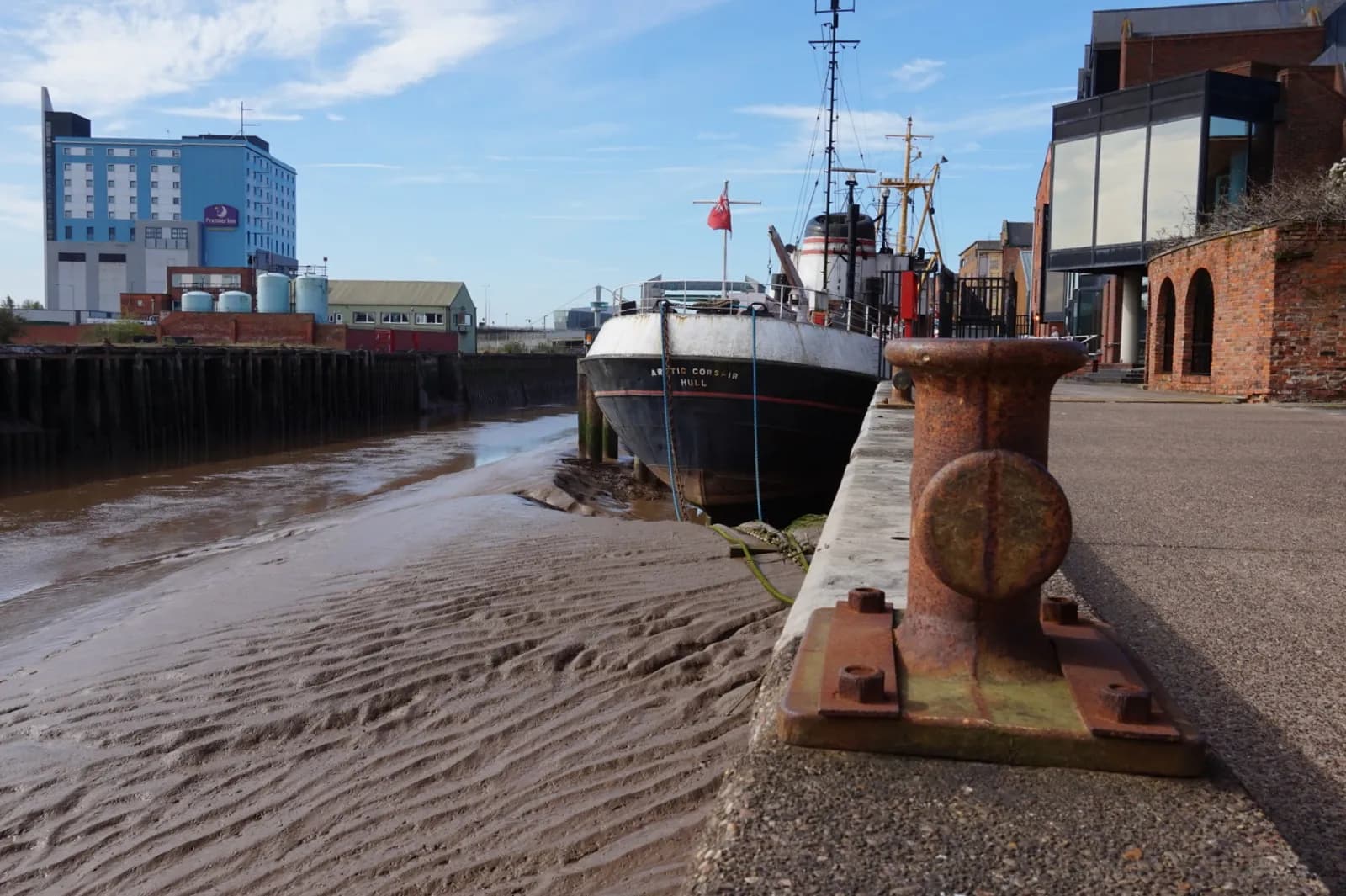 A historic black and white ship is docked on a muddy riverbank next to a concrete pier with a rusted bollard.