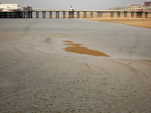 A long pier extends from the left across a sandy beach, with calm water in the foreground and a town visible in the background.