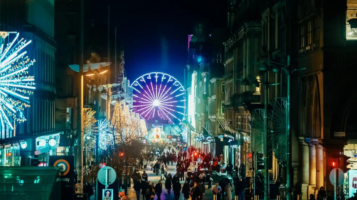 A Ferris wheel illuminated in purple and white lights towers over a busy street at night, lined with buildings decorated with sparkling holiday lights.