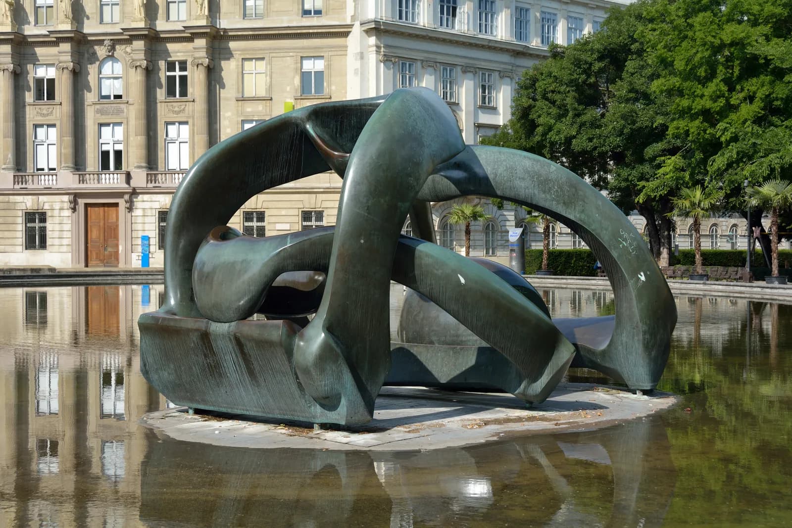 A large, abstract bronze sculpture with flowing, interconnected forms sits in the center of a reflective pool, with an ornate building and green trees in the background.