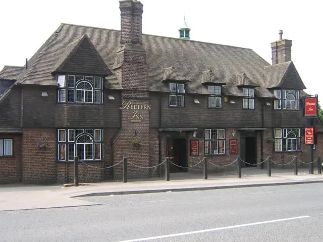 A large, brick building with a dark shingled roof, featuring multiple bay windows and prominent brick chimneys, identified by a sign as "Redfern Inn" with a street and sidewalk in the foreground.