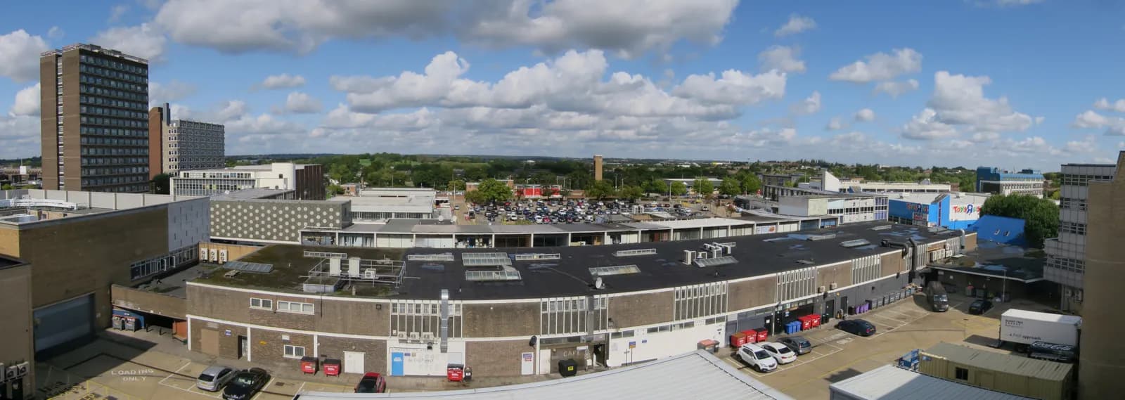 A wide shot of a town with several buildings, a parking lot, and a distant green landscape under a partly cloudy sky.