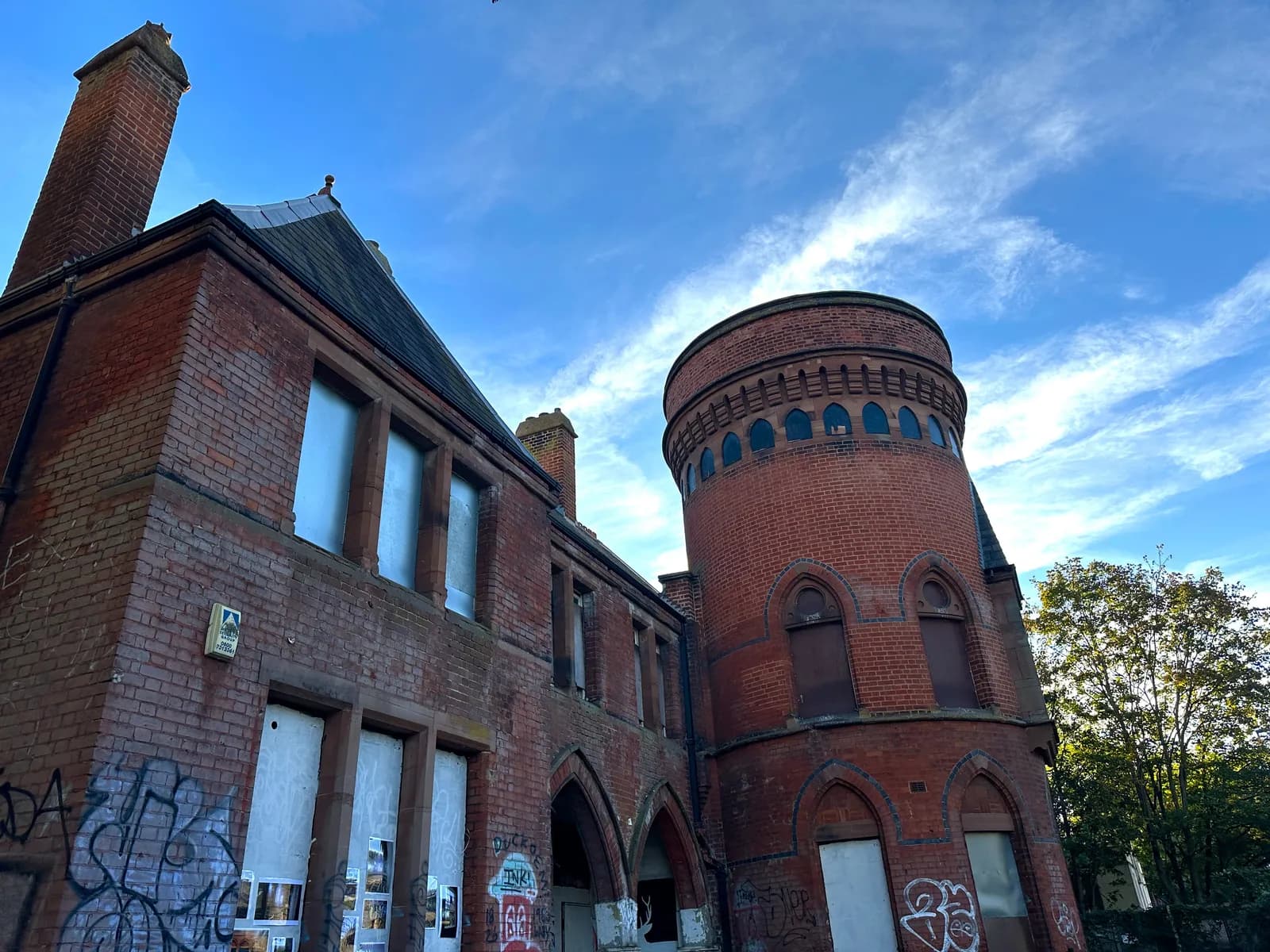 A historic red brick building with a round tower and boarded-up windows, covered in graffiti, stands against a bright blue sky with wispy clouds.