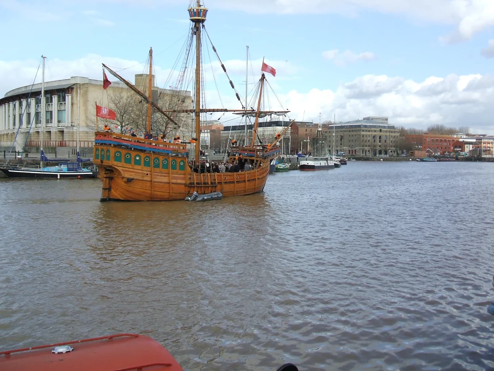 A wooden tall ship with masts and sails, flying red flags, floats on a murky river next to a city with large buildings and other boats.