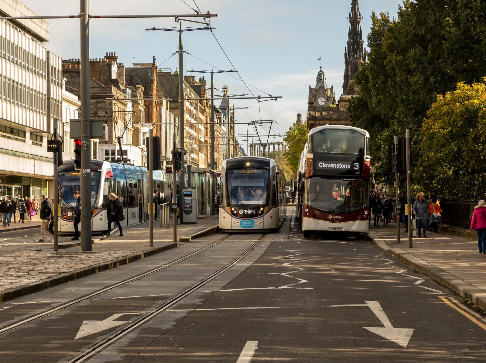 A street scene with two trams on the left, a double-decker bus on the right, people walking on the sidewalks, and buildings in the background.