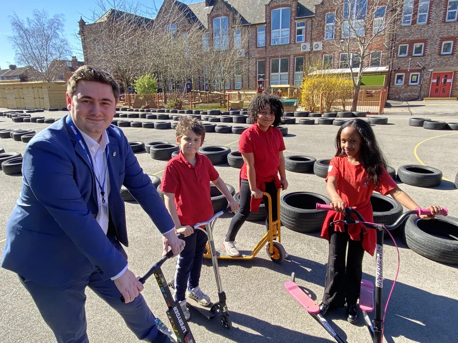 A man in a blue suit and three children on scooters pose in a schoolyard with tires arranged on the ground and a building in the background.