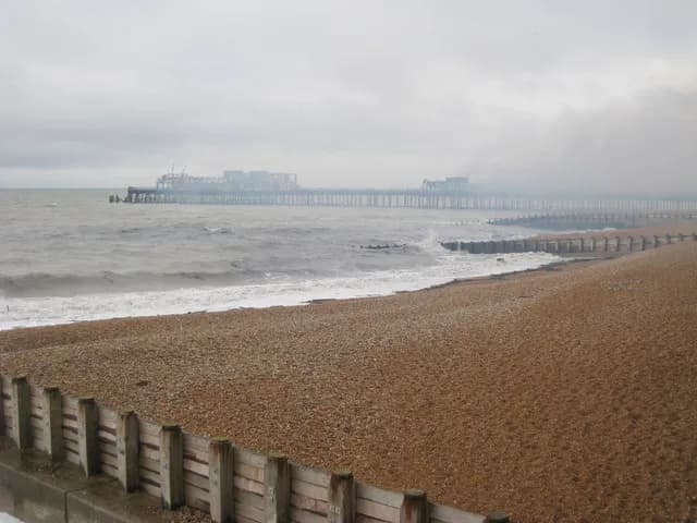A long pier stretches into the sea under a grey, cloudy sky, with waves crashing against a pebble beach and wooden groynes in the foreground.