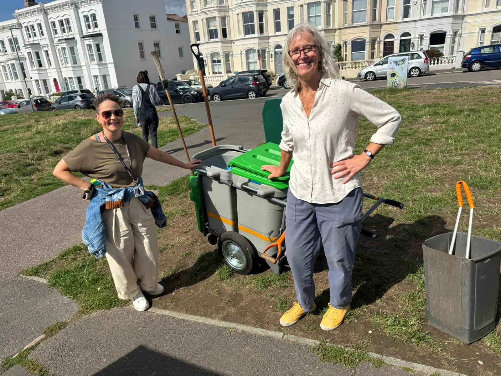 Two women, one with gray hair and glasses, stand smiling next to a gray and green cleaning cart and a rubbish bin, with buildings and cars in the background.