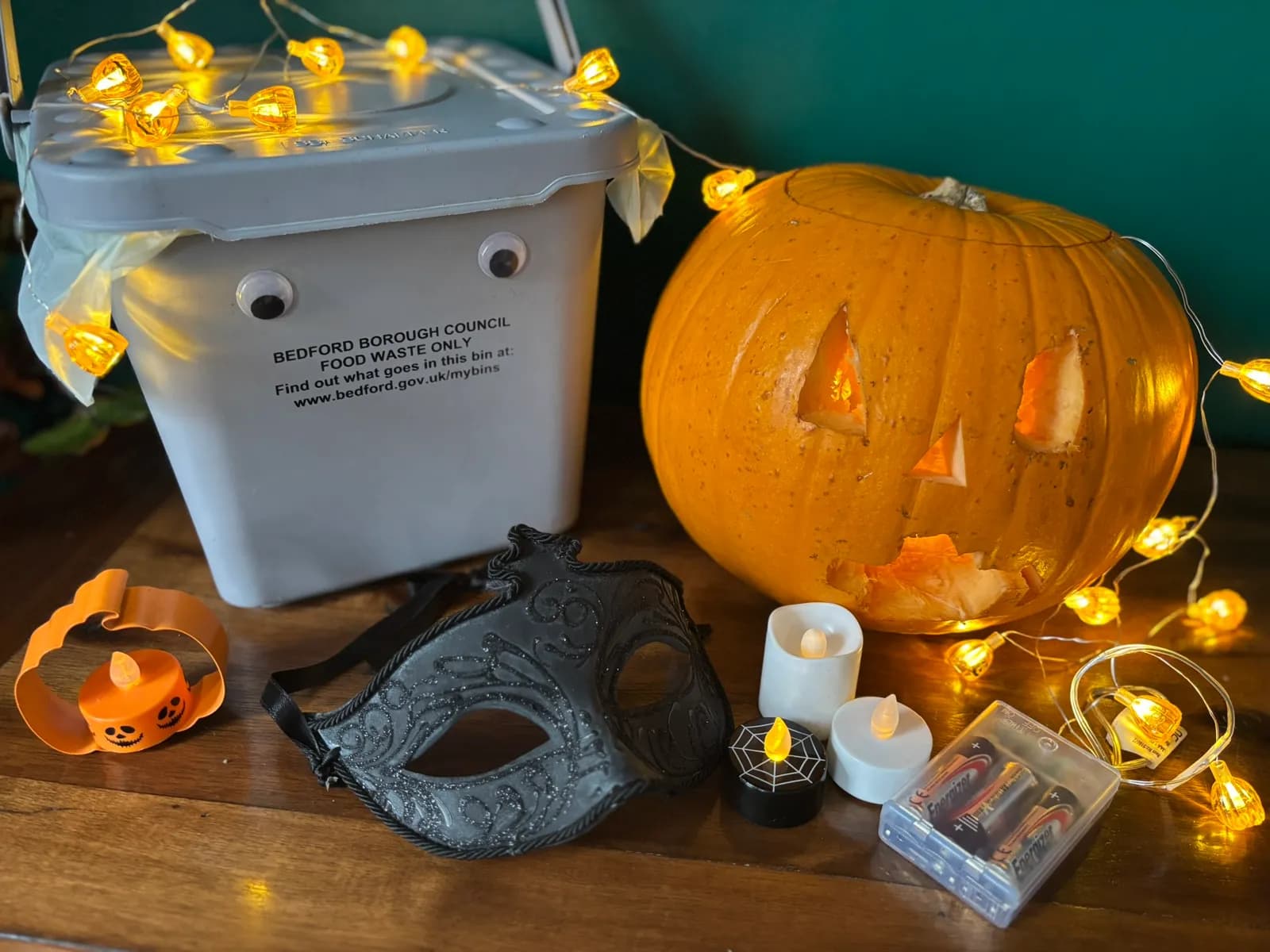 A Halloween-themed display featuring a carved pumpkin, a grey food waste bin with googly eyes, a black masquerade mask, and several LED candles and string lights, all arranged on a wooden surface.